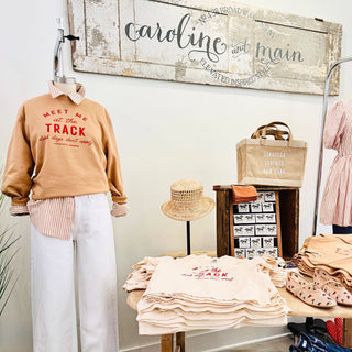 Display of clothing and accessories with 'Caroline and Main' branding in the background featuring the beige sweatshirt with red text 'Meet Me at the Track - Dark Days Don't Applu text. 
- Saratoga Springs, NY Boutique